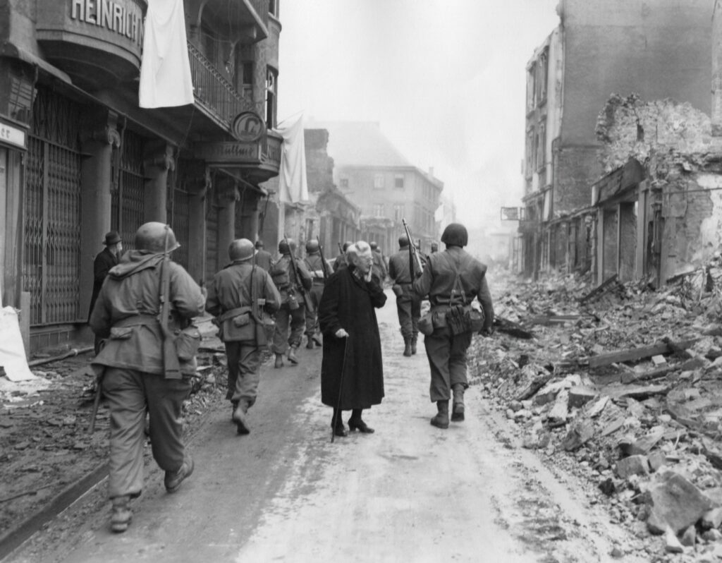 Photo of town ruins and military men walking with rifles with old woman in the street