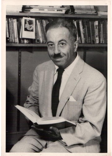 Man in black and white photo smiling holding book while wearing suit jacket and tie