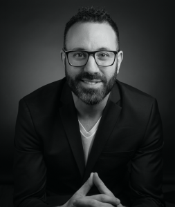 A black and white picture of Danny M. Cohen. He is wearing a black blazer and white t-shirt, and leaning slightly forward, with his shoulders rounded. His fingers are steepled and he is smiling. He is wearing glasses and has a short beard. 