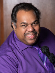 A front-facing picture of Daryl Davis, an African American man in a three-fourths pose wearing a purple shirt with a purple tie. A microphone is in the foreground, and he sits against a brown background.