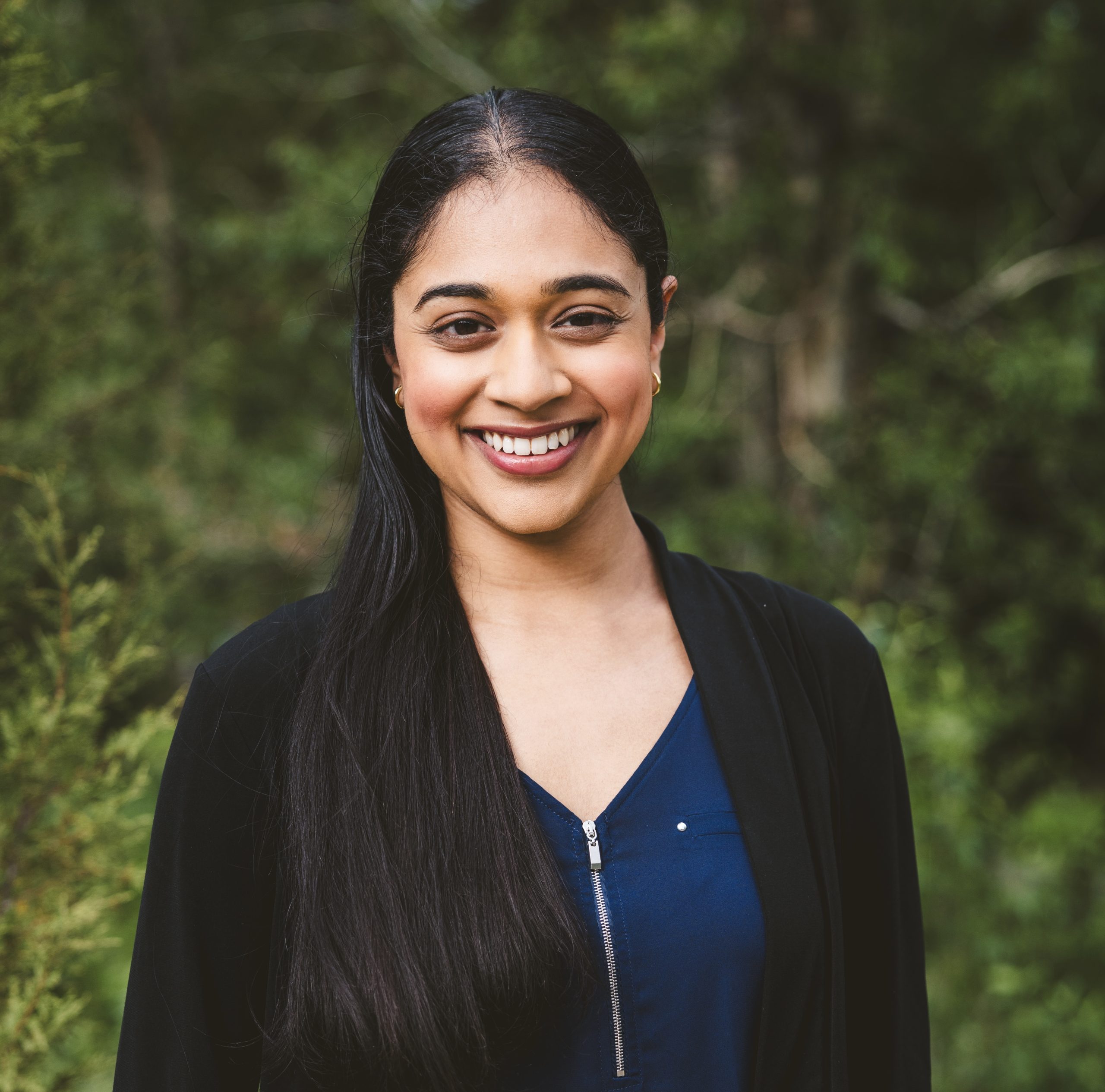 A headshot of Trisha Prabhu. She is wearing a black blazer and a navy blue shell. Her hair is tucked over one of her shoulders, and she is standing in front of a nature background. She is smiling.